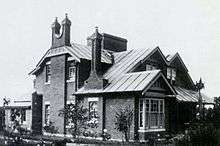 Black and white photograph of the facade of the brick house. Two storeys with a two-pronged chimney rising up the front wall.