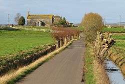 Stone building at the end of narrow lane with water filled ditches on either side. Surrounded by fields and trees.