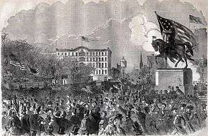 Crowd surrounding an equestrian statue topped by a huge U.S. flag