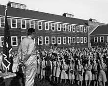 A large crowd of men and women in uniform listens to a fat man in uniform speaking at a microphone. They are wearing the Army Service Forces sleeve patch. The women are at the front and the men at the back. Beside him is the flag of the Army Corps of Engineers. Behind them are wooden two-storey buildings.