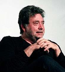 A photo of a bearded, black haired Mexican male with sitting with his hands clasped over his knee. He is wearing a black shirt and black pants.