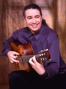 Laucke, seated in half profile, playing his guitar and smiling while looking away from the camera