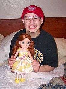 A young woman sitting upright in a bed, her legs under the blanket and the headboard and pillows behind her. She holds a large doll in front of her and is wearing a T-shirt, large glasses and a red baseball cap while smiling broadly