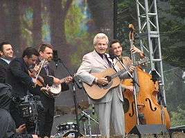 A man wearing a blue dress shirt, standing behind a microphone stand and holding a guitar.