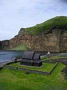 Picture of a small wooden church by an inlet of the sea, with mountains in the background.