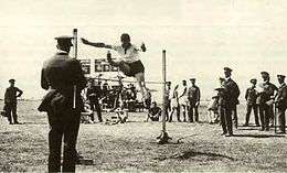 High jumper watched by a crowd of people, most wearing dark military uniforms with peaked caps