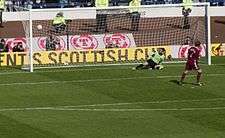 A football player scores a goal against the opposing goalkeeper from a penalty-kick. Stewards and camera-operators are visible behind the goal net.