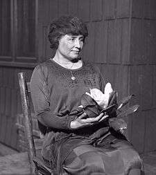 A woman with full dark hair and wearing a long dark dress, her face in partial profile, sits in a simple wooden chair. A locket hangs from a slender chain around her neck; in her hands is a magnolia, its large white flower surrounded by dark leaves.