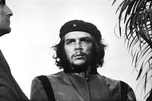 Black-and-white image of a man looking to his right. He wears a black beret with a little "x" and a closed jacket. His hair is shoulder-length, curly and black. On his right there is a man looking to him and on his left, there is a plant.