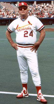 White man in his mid fifties wearing a white baseball jersey with red trim, a matching cap and sunglasses, standing on a baseball field