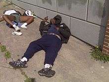 A photograph of a man and a woman lying on a sidewalk, the woman wearing one white shoe while its match lies beside her, the man wearing two shoes