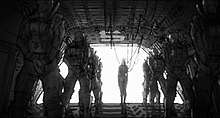 Black-and-white drawing of a group of large, suited soldiers and a small woman connected to wires in front of an open door