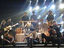 Madonna sitting onstage singing, surrounded by her dancers in a circle.