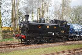 A pannier tank locomotive is pulling a milk wagon. The locomotive is painted black. The words "GREAT WESTERN" are shown in yellow with red shading on the side of the pannier tank. The front buffer beam is painted red.