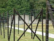 Taken from outside the reconstruction of a barracks, the photo shows a barbed wirefence, and beyond it a grassy area with a small timber hut