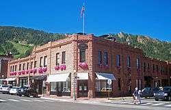 A two-story light brown stone building on a street corner, with a narrow entrance facing the camera in the center of the image. It is lit by the sun from the left. There are storefronts along both sides at street level; all the windows on the second story . Above the main entrance is an awning with the word "Dior" on it, and above the second-story window a semicircular black plaque with "Brand Building, 1891, Aspen, Colorado" on it in gold letters. An American flag is on a pole above that. In the background is a rocky and forested ridgeline with some cleared areas.