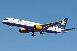 A mostly white Boeing 757 with blue and yellow trim preparing for landing against a blue sky. Landing gear and flaps are fully extended in  final approach configuration.