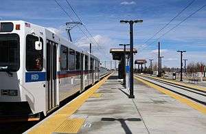 A white train is parked at a recessed side platformed station with canopies and signage visible.