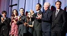 A man in a black suit, a woman in a pink dress, a man in a plaid suit, a woman in a black dress, a Japanese man in a black suit, and an old man in a blue suit clap their hands, while a man in a black suit stands. A microphone stand is in the foreground, and blue curtains are in the background.