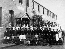 Posed, group photo of students and teachers, dressed in black and white, outside a brick building in Regina, Saskatchewan in 1908