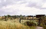 A hillside with multiple barbed-wire fences running parallel to each other, with fruit trees, a barn and a watchtower in the background.