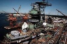 In the background a large shipyard can be seen, with cranes and dockyard warehouses. In the foreground a massive steel superstructure can be seen, decked out in scaffolding with tarps covering sensitive equipment and wires across the superstructure in all directions.