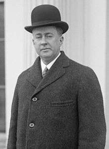 Black and white image of a clean shaven man with drooping eyelids, aged about forty. He is wearing a black bowler hat, white shirt, tie and dark overcoat.