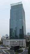 Ground-level view of a blue, glass high-rise; the tower sits behind a small, white, stone, window-dotted facade