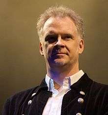 A Caucasian male with silver cropped hair and a goatee looking off camera. He is wearing a white shirt opened at the neck with black jacket.