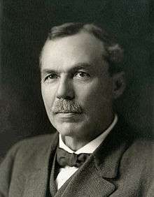 A photo of the head and shoulders of a man wearing a three piece suit and bow tie. In his forties, his hairline is partially receded and one eyebrow is noticeably higher than the other.