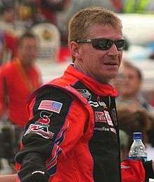 A man in his forties wearing black sunglasses and an orange-and-black jacket with sponsors logos