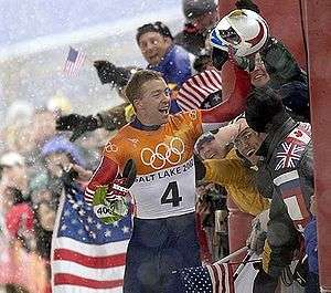 A man celebrates near a joyful crowd waving American flags behind a security fence. Holding a helmet high in his :) left hand, he wears a blue, red, white, and orange jumpsuit with the Olympic rings, the words Salt Lake City, and a number four in the chest.