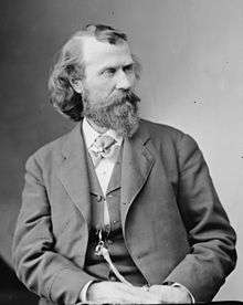 A finely detailed monochrome photograph portrait of a bearded and mustachioed man in his 30s or 40s, shown from the waist up, wearing a jacket and vest over a white shirt with its collar closed by a cravat secured by a jeweled finger ring, a multi-corded watch fob hanging from a vest button, decorated by another ring, the man's hands together in his lap, his body leaning to the left and the head turned to the right, his dark hair full and long in the back, long but thin on top, revealing a high forehead