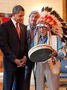 Joe Medicine Crow in full feathered headdress plays a drum with a man in a suit watching