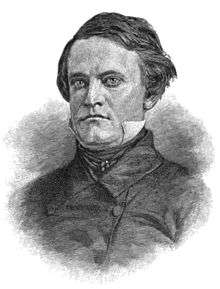 A man with thick, dark hair wearing a high-collared white shirt under a black jacket and tie