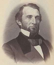 A man with black hair and a black beard wearing a black jacket and tie and white shirt