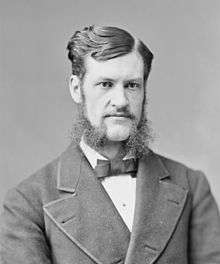 A man with dark, wavy hair and bushy sideburns wearing a black jacket and bowtie and white shirt