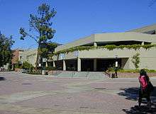 A photograph of a gray, three-story building on campus. The second story is offset back from the first and the third is offset form the second. The building has plants hanging over the second story onto the front of it. It is decorated with three unreadable banners. There are eight steps that lead to its covered entrance.