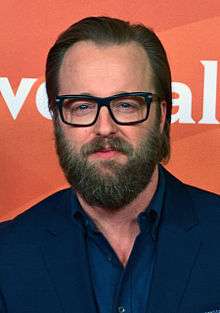 A photograph of a smiling middle-aged Caucasian man with thick beard and combed hair, wearing glasses and a dark blue suit and shirt.