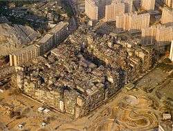 A large solid block of ramshackle buildings varying in height, with many taller buildings and some mountains in the background.