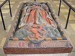 A stone tomb, with the effigy of a robed man on top, with a coat of arms at his feet displaying a red panther on a blue-and-gold striped background.