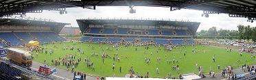  A view of a football stadium with a stand opposite and to the left, and an open space to the right. There are people on the pitch