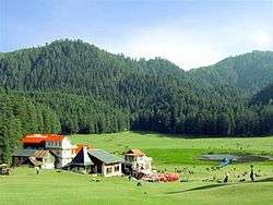 Daytime sunlit view of a verdant bowl-shaped meadow with a small pond in a valley rimmed by several prominent hills covered in evergreen forest. At centre-left and looking out on the meadow, a small cluster of tourist lodges with pitched roofs, one a bright red. Around them and the pond are small, gaily coloured gazebos and shade-giving umbrellas. Several dozen tourists and sheep mill about the pond and in the meadow.