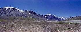 Some tall, blue-grey mountains rise out of brown soil below a deep blue sky