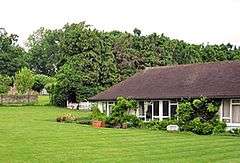 A white building with a black roof beside a large, well-kept lawn. Many trees are in the background of the picture, behind the building.