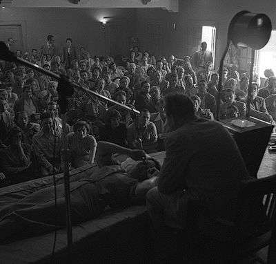 A mostly seated crowd watches as Hubbard, seated on a chair, speaks to a woman lying prone in front of him.