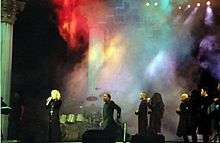 Faraway image of Madonna wearing a black robe with a crucifix and singing to a microphone she holds in her hand. The staged is filled with a colored smoke and some drums and a cross can be seen.