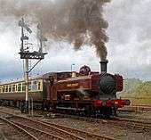 A pannier tank locomotive is passing a semaphore signal, leading a train which includes one visible passenger carriage. The pannier tanker is maroon, apart from the black chimney, brass safety valve cover,  red buffer beam, and grey coupling rods. The tank, cab, splashers and toolbox are all lined in yellow. London Transport is written on the side of the tank, and L.94 on the side of the cab, again both in yellow. A plume of brown smoke comes from the chimney, and there is as whisp of steam from under the pannier tank. The passenger carriage is cream and brown with a grey roof.