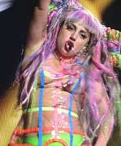 A closeup of Gaga, in a neon colored dress with her head tilted to the left side. She has her right hand raised and is singing to her mouthpiece.