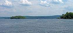 Image of a boat moving away from the photographer on a wide lake with two forested islands visible in the foreground. Mountains rise in the background.